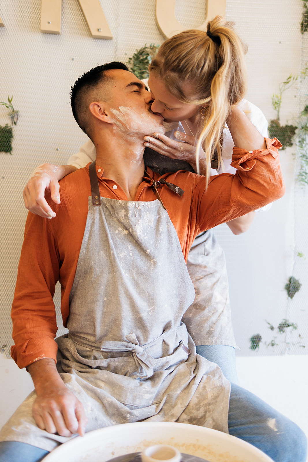 a woman kissing a man as he makes pottery during date night photoshoot 