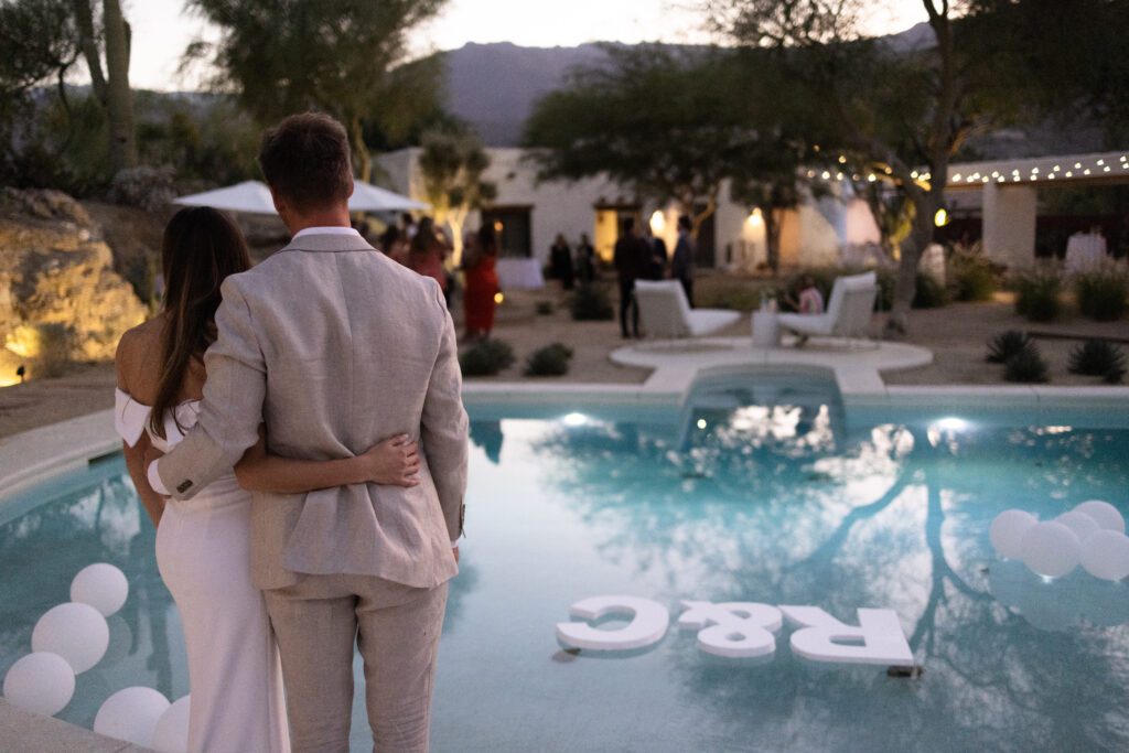 bride and groom standing together by the pool looking at their guests 