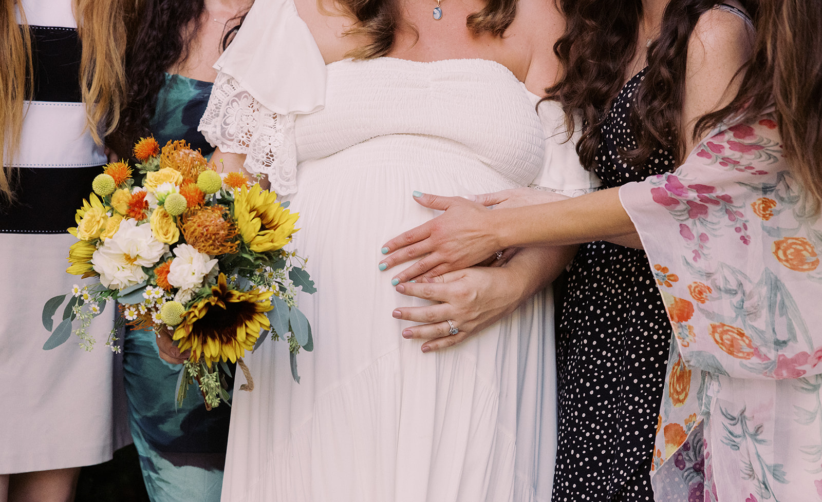 the bride standing with her friends as they place a hand on her belly 