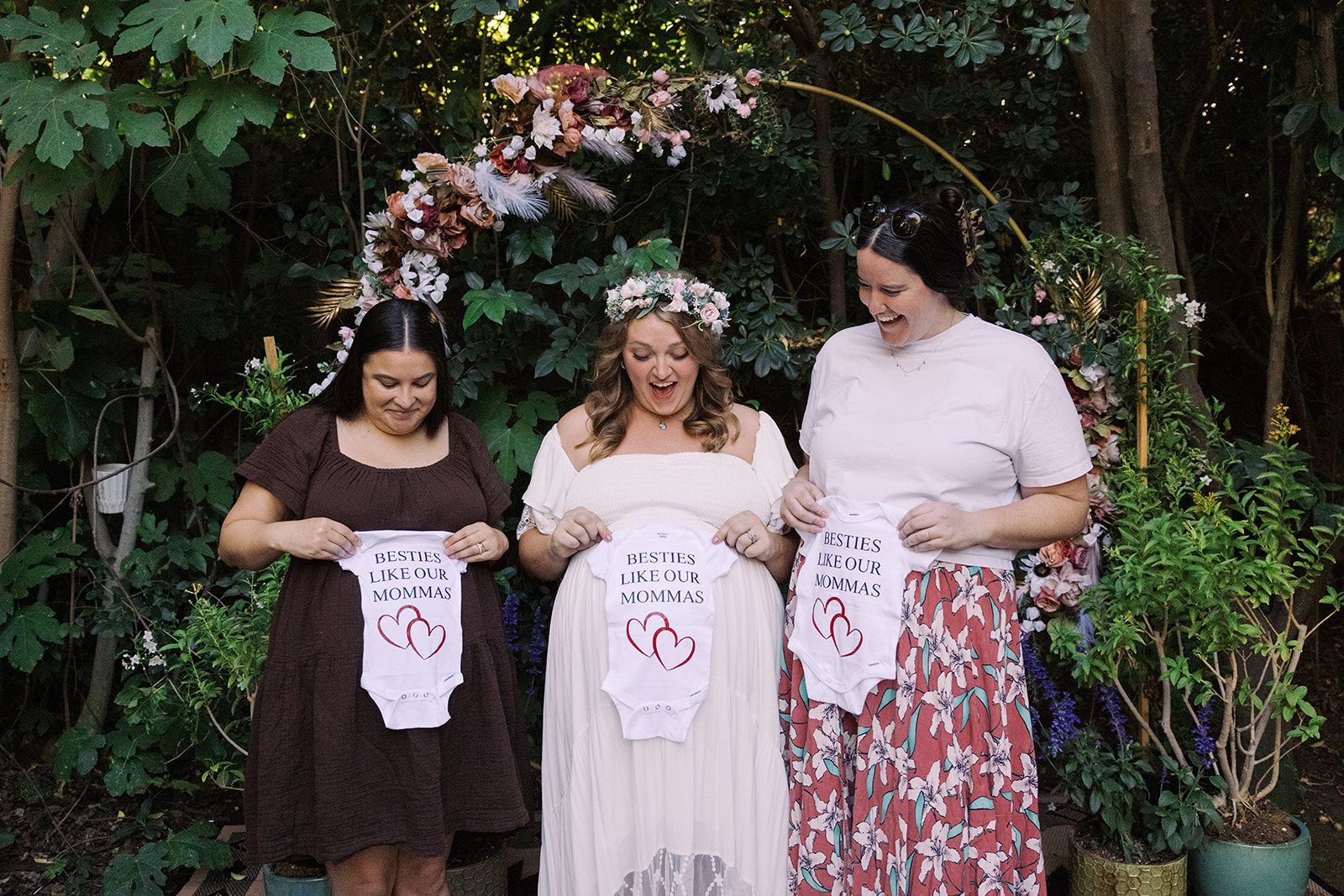 the bride and two pregnant friend holding up onesies that say 'besties like our mommas'