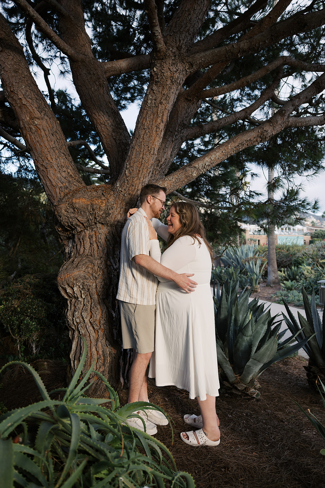 a couple laughing as they stand by a tree during their Orange County engagement photos 