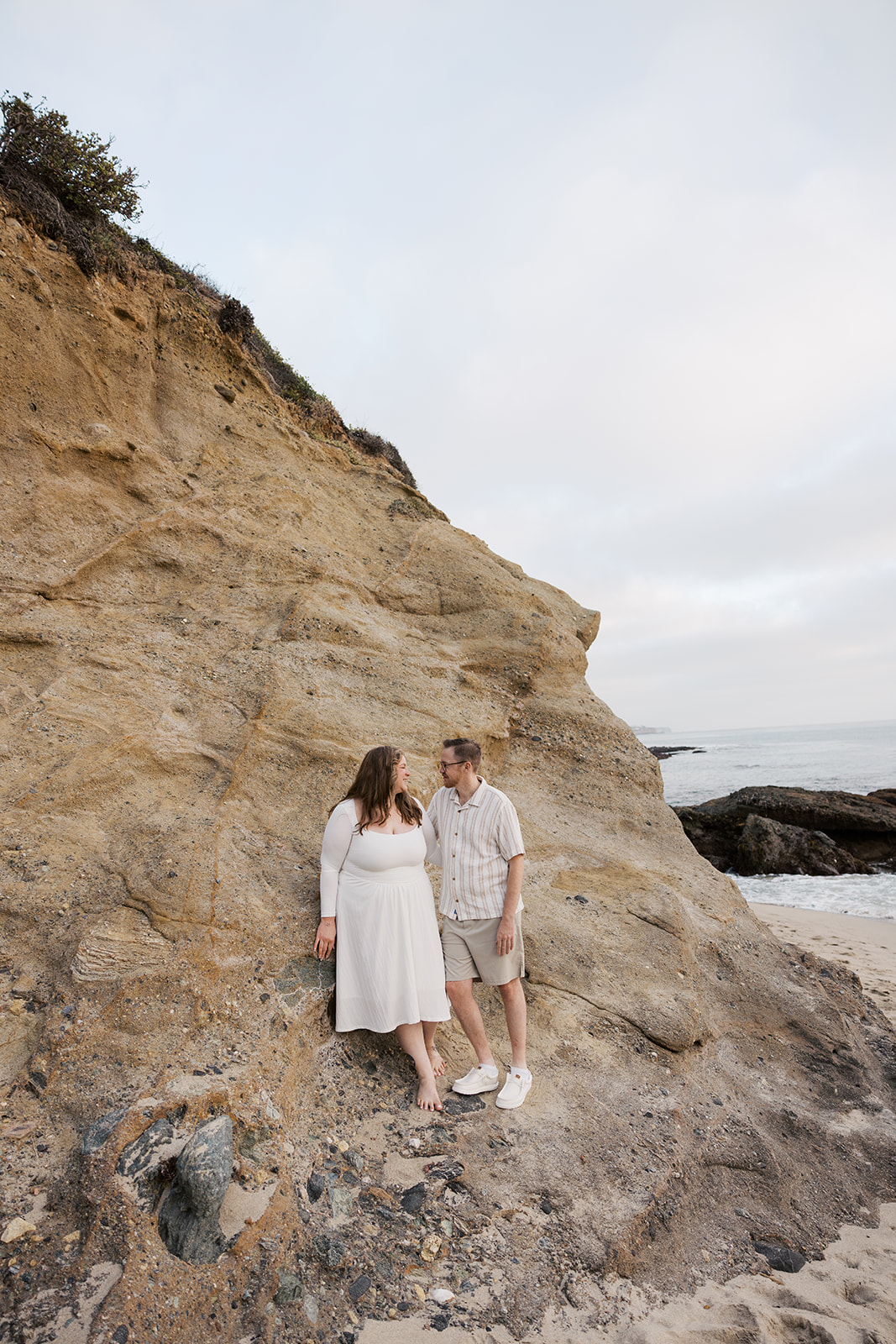 a couple standing by a rock cliff at the beach during their beach engagement photos in California